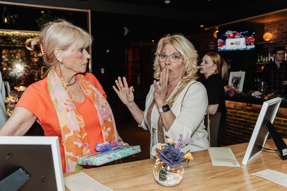 Two women chatting at a bar counter, one in orange floral top, other in white jacket with pearls, looking surprised.