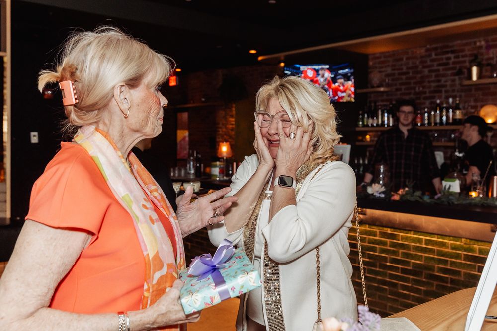 Two women sharing a surprise moment at a bar, one presenting a gift while the other reacts with joy.