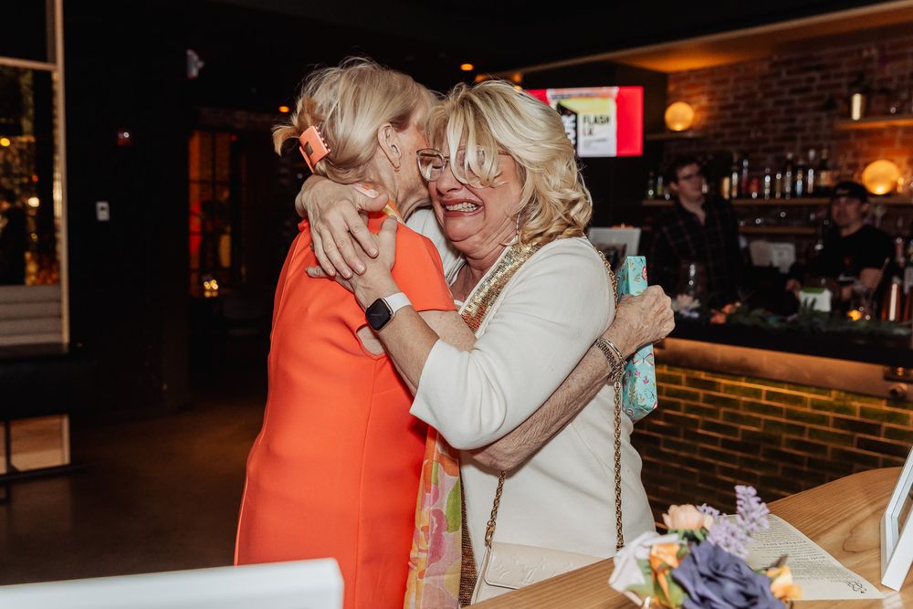 Two blonde women sharing a warm embrace at a wedding reception near a bar with brick walls.