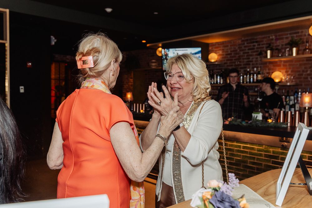 Two women in elegant attire engage in lively conversation at a stylish bar or restaurant event.