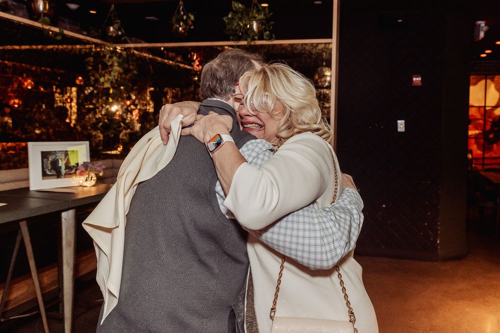 Two people share a warm embrace at an indoor evening event with ambient lighting.