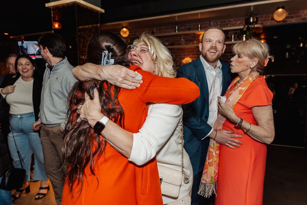 Two women in orange and white dresses share a warm hug at a festive indoor gathering with guests nearby.