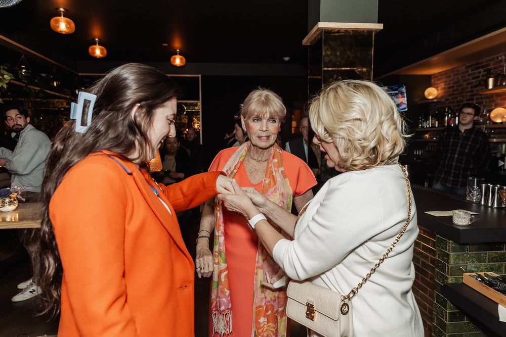 Three women in colorful attire socializing at an upscale restaurant event, with guests mingling in the background.