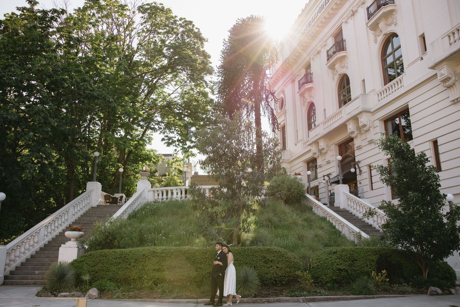 Couple embracing in front of elegant white European building with grand staircases and lush green trees in golden sunlight.