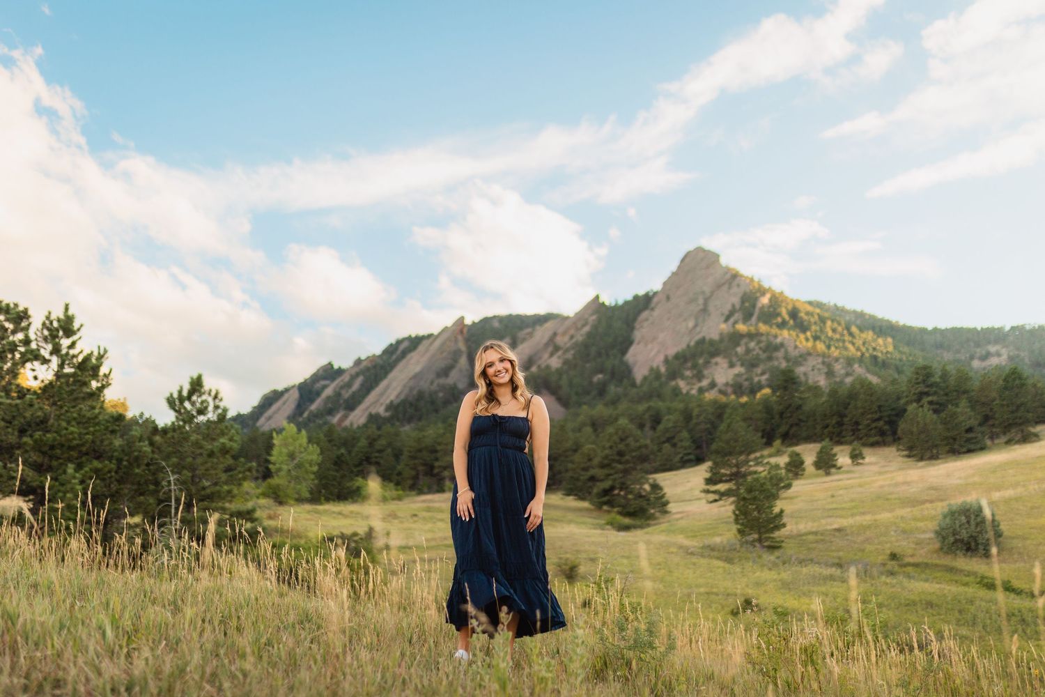 A high schooler in a navy blue dress stands in a meadow with the iconic Flatiron Mountains of Boulder, Colorado in the background.