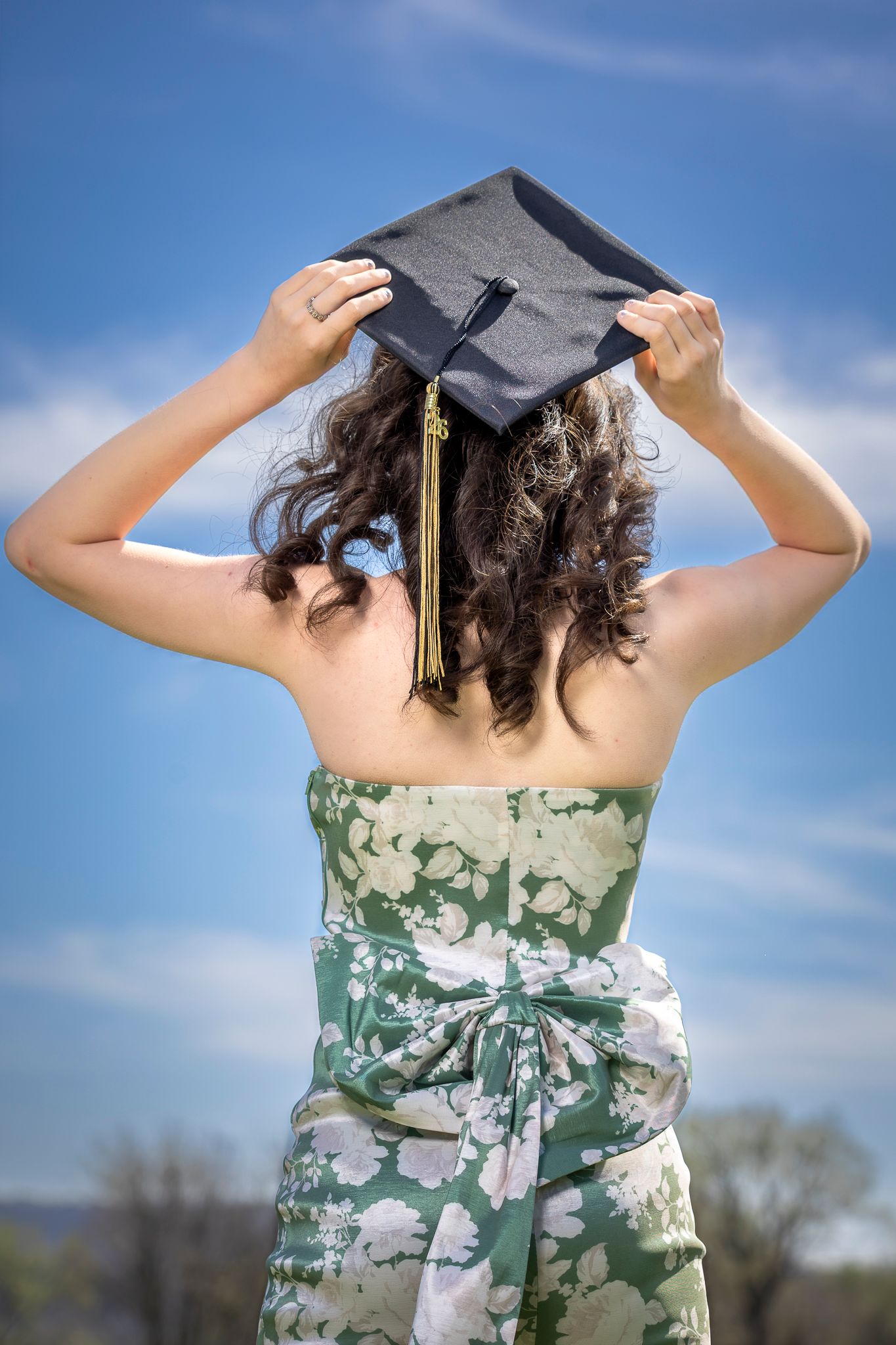 Graduate in floral green and white strapless dress with flowing sash poses outdoors in graduation cap.