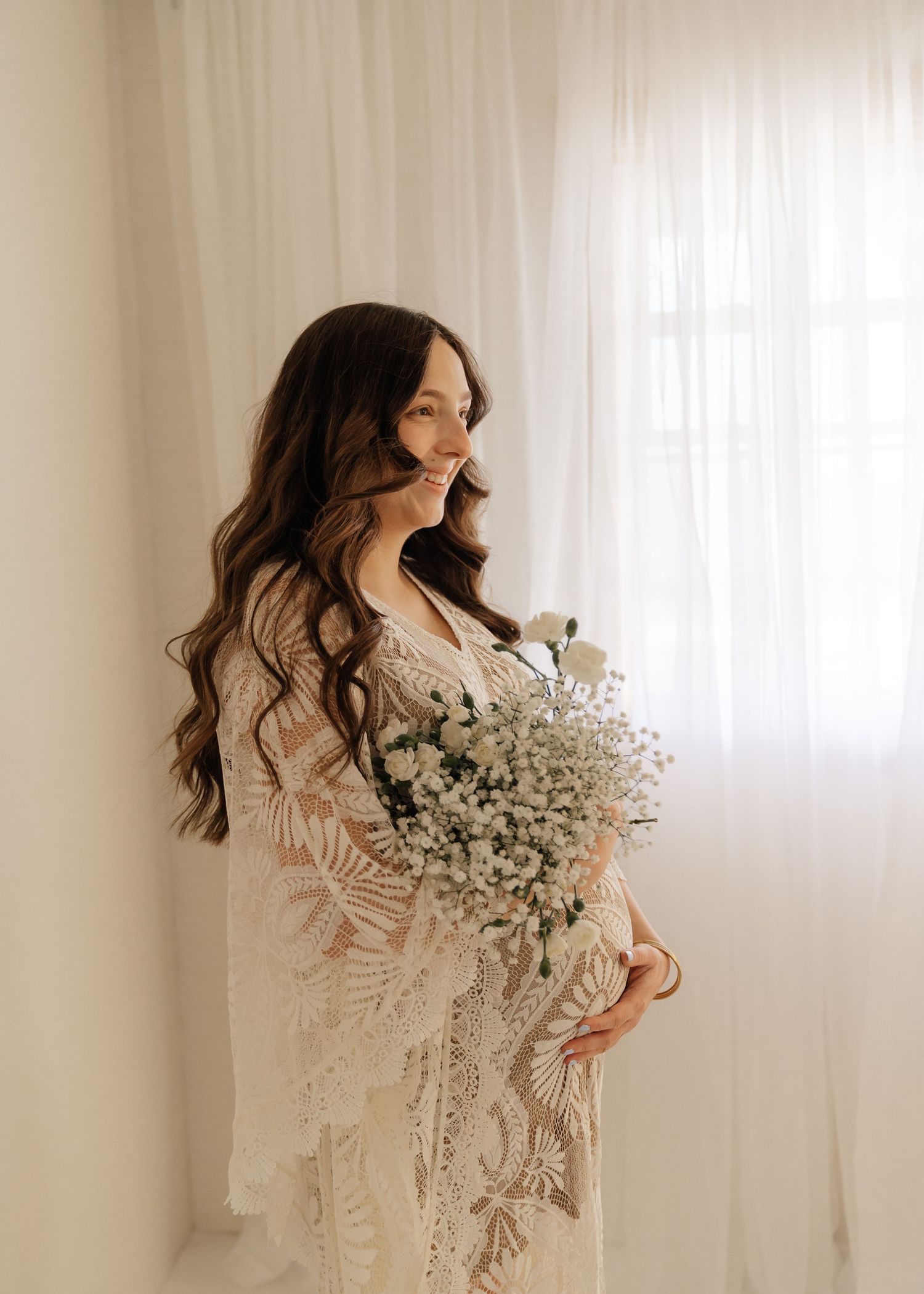 Woman in white lace dress holds baby's breath flowers while posing against light curtained window.