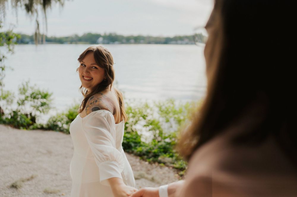 A bride in a white dress sits on a sandy beach near the water during a romantic photoshoot.