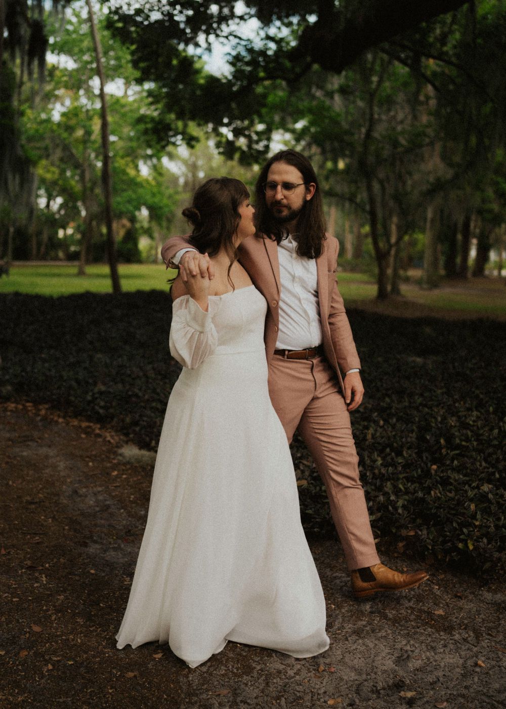 Two people in coordinating wedding attire share intimate moments while walking through a wooded outdoor setting.