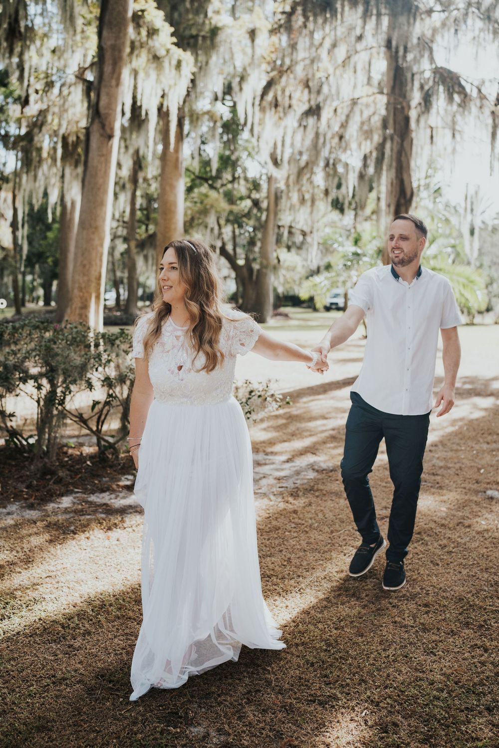 A couple in white and denim hold hands while walking through a sunlit forest with Spanish moss hanging overhead.