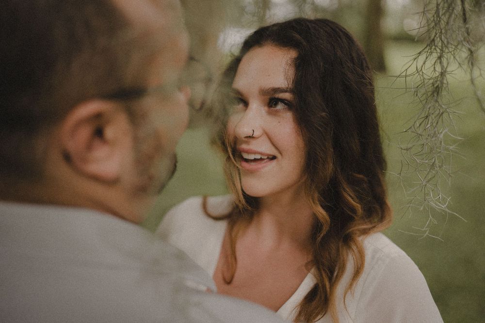 A couple shares a tender moment outdoors with soft natural lighting and hanging foliage in the background.