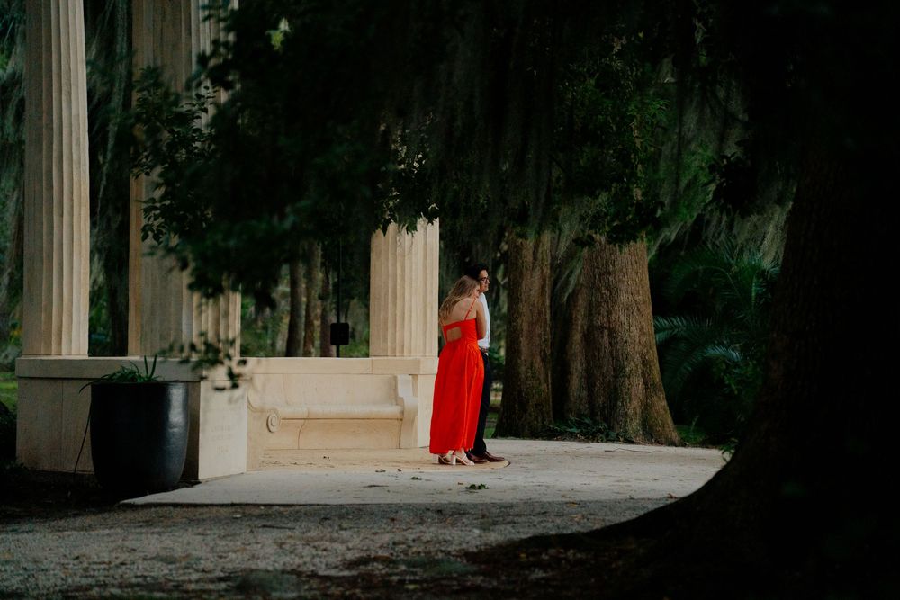 A person in a red dress walks along a colonnade pathway surrounded by lush greenery and stone pillars.