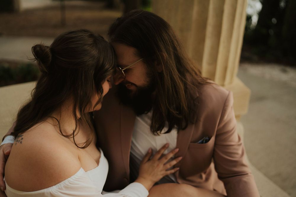 A romantic couple shares an intimate moment together outdoors, with one wearing a pink blazer and the other in white.