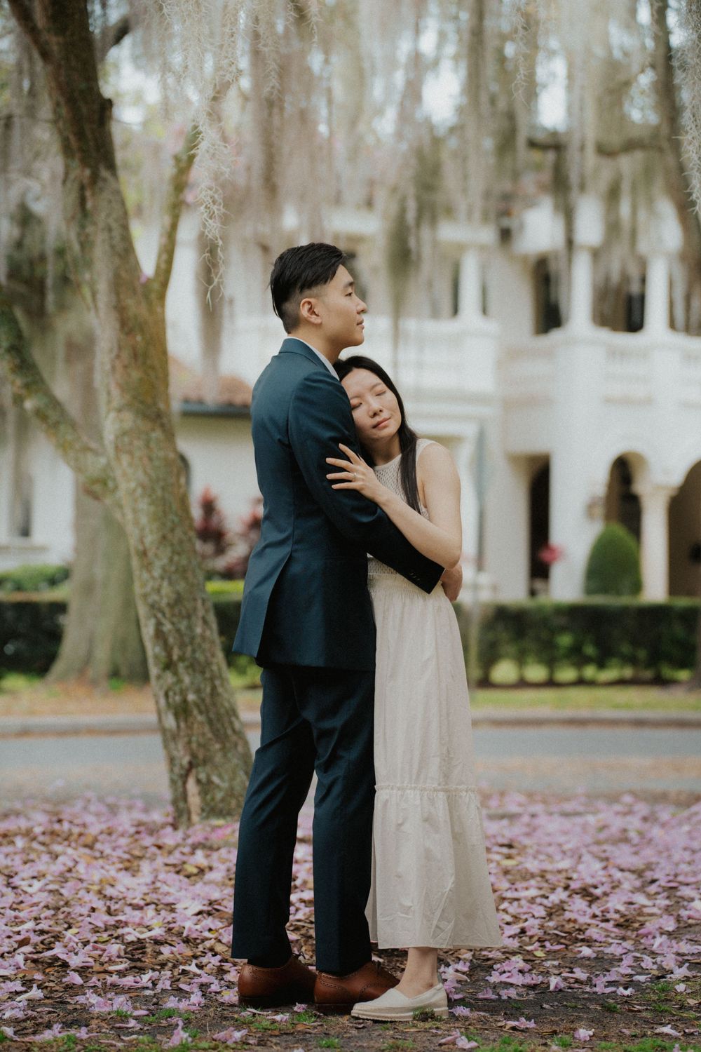 A romantic couple embraces under blooming trees with pink petals scattered on the ground at a stately venue.