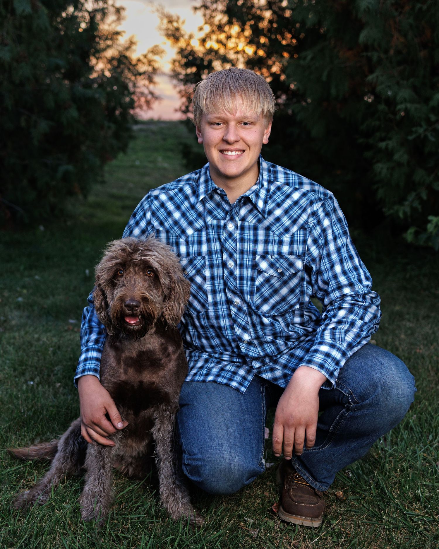 A senior boy in a blue plaid shirt kneels next to a chocolate-colored labradoodle dog on grass at sunset.