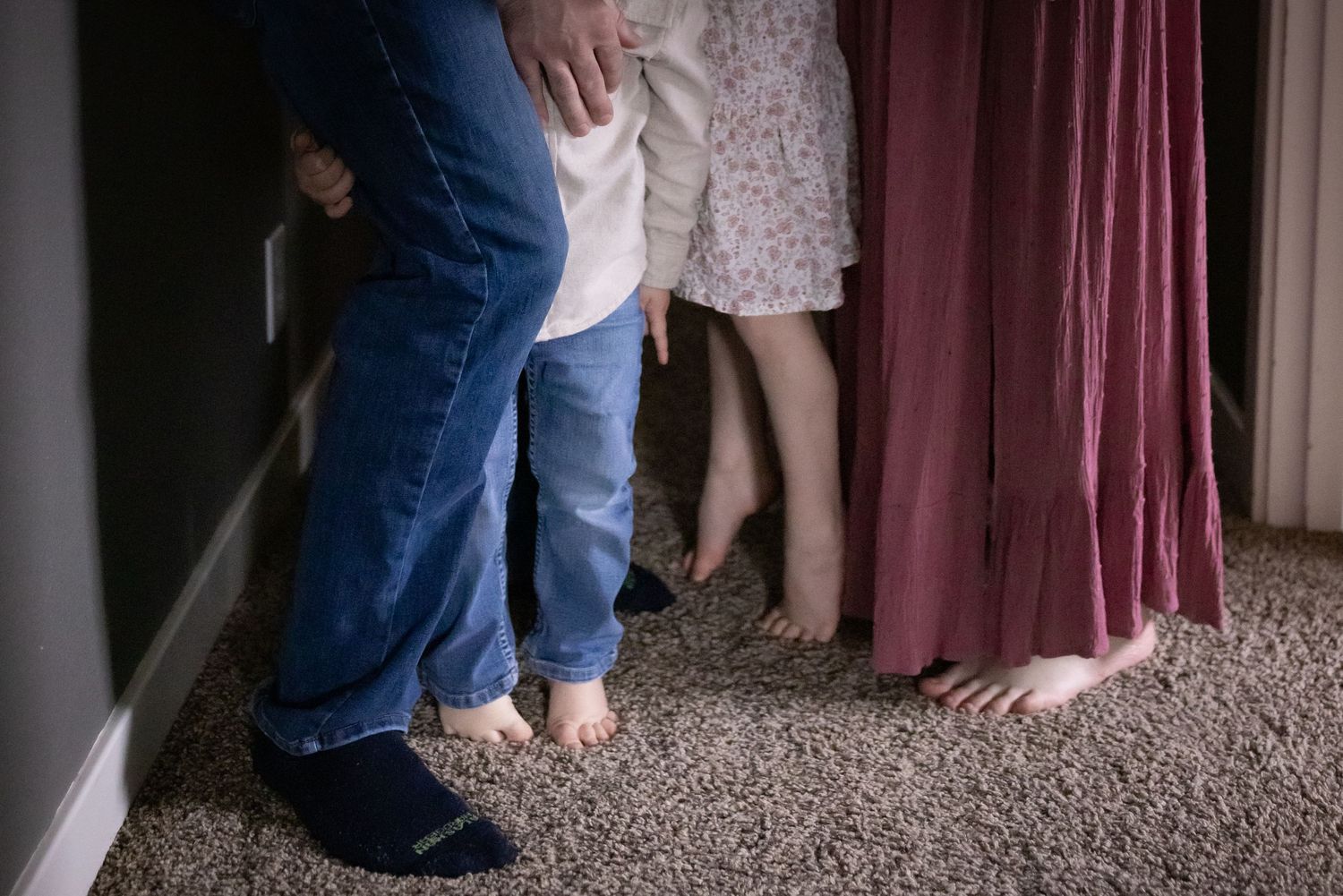 Family standing barefoot together indoors, showing legs and feet on carpet.