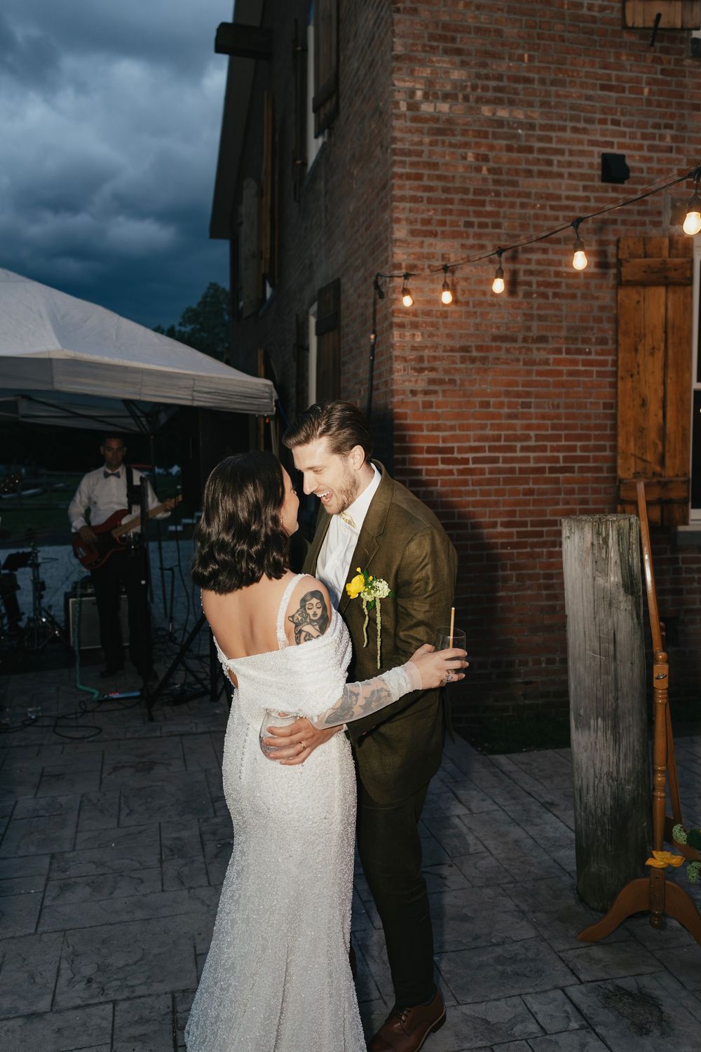 Newlyweds dance together on stone patio outside brick building with string lights overhead.