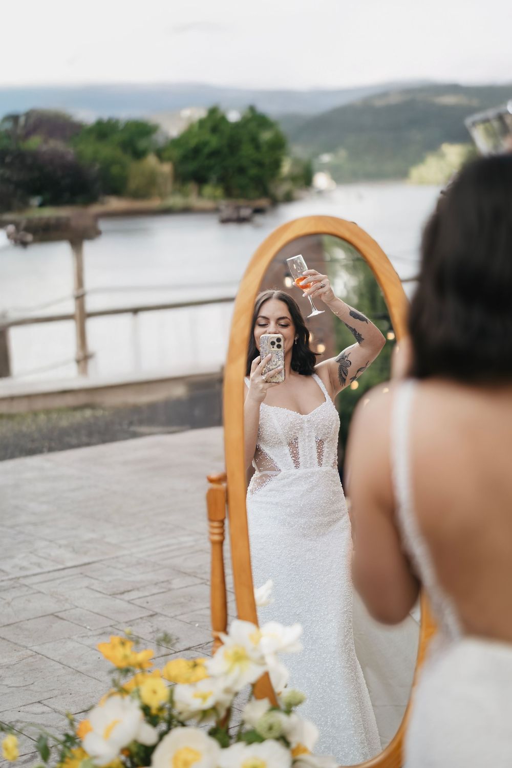 A bride takes a selfie in an ornate wooden mirror outdoors with water and mountains in the background.