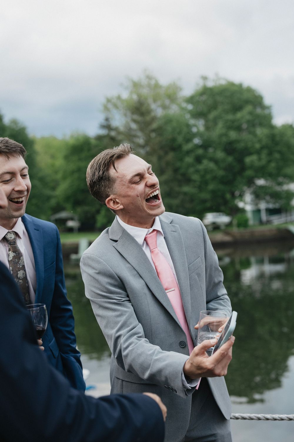 Groomsmen laughing together by a lake during wedding photos wearing gray and navy suits with pink ties.