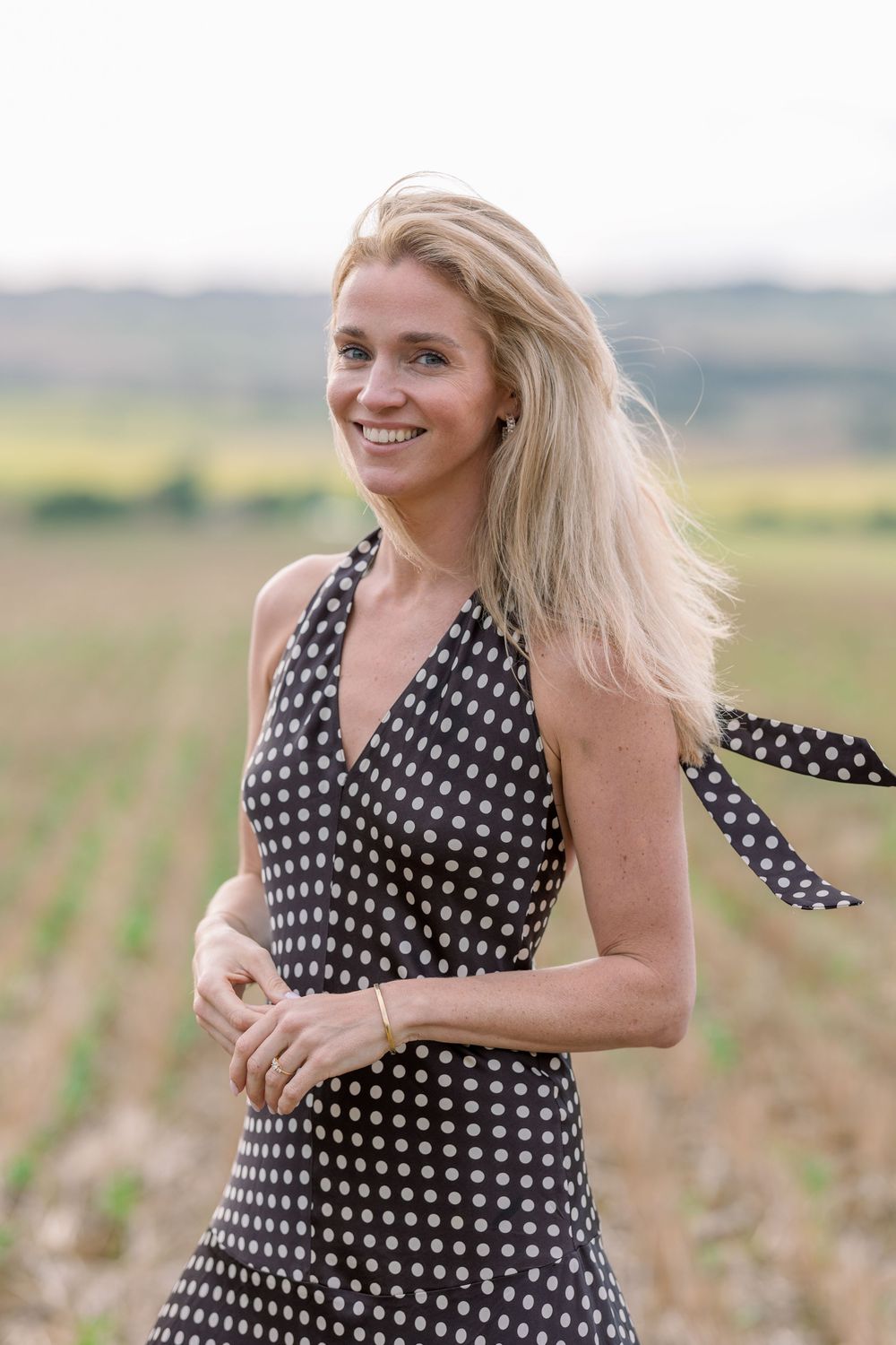Woman in polka dot dress poses playfully in a sunny outdoor field setting.