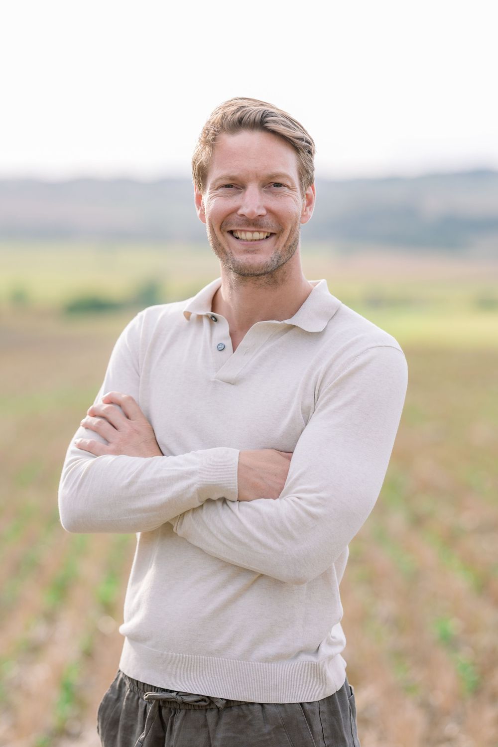 Person in white sweater smiles warmly while posing against a natural outdoor backdrop.
