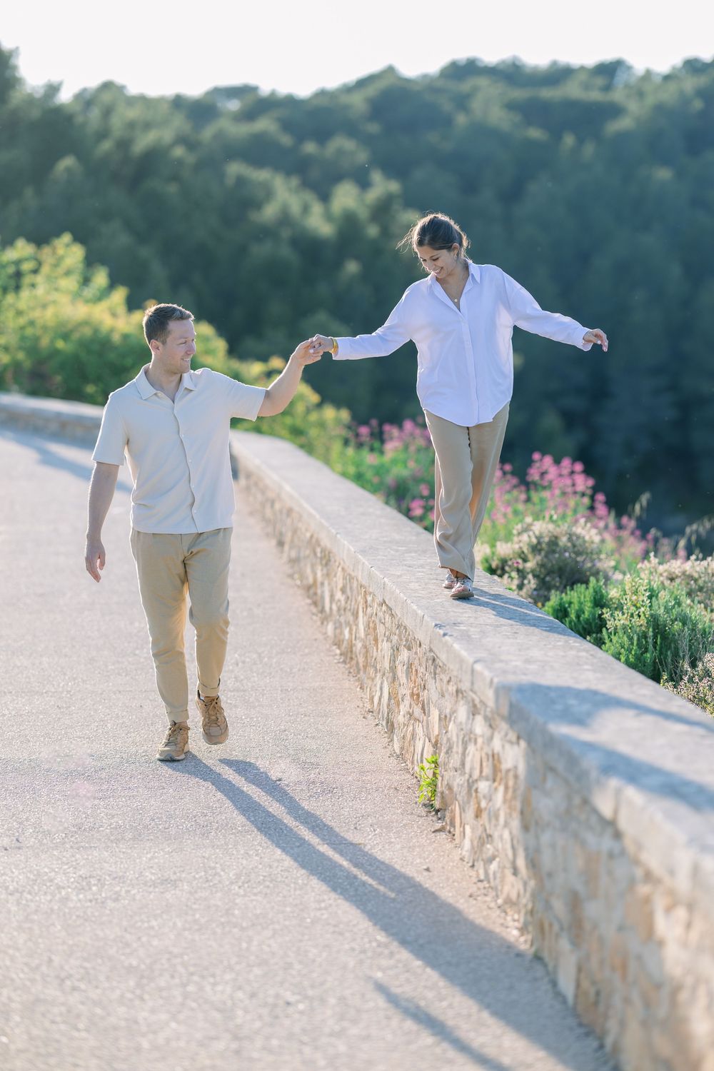 Two people walk playfully along stone wall path with flowers and greenery.