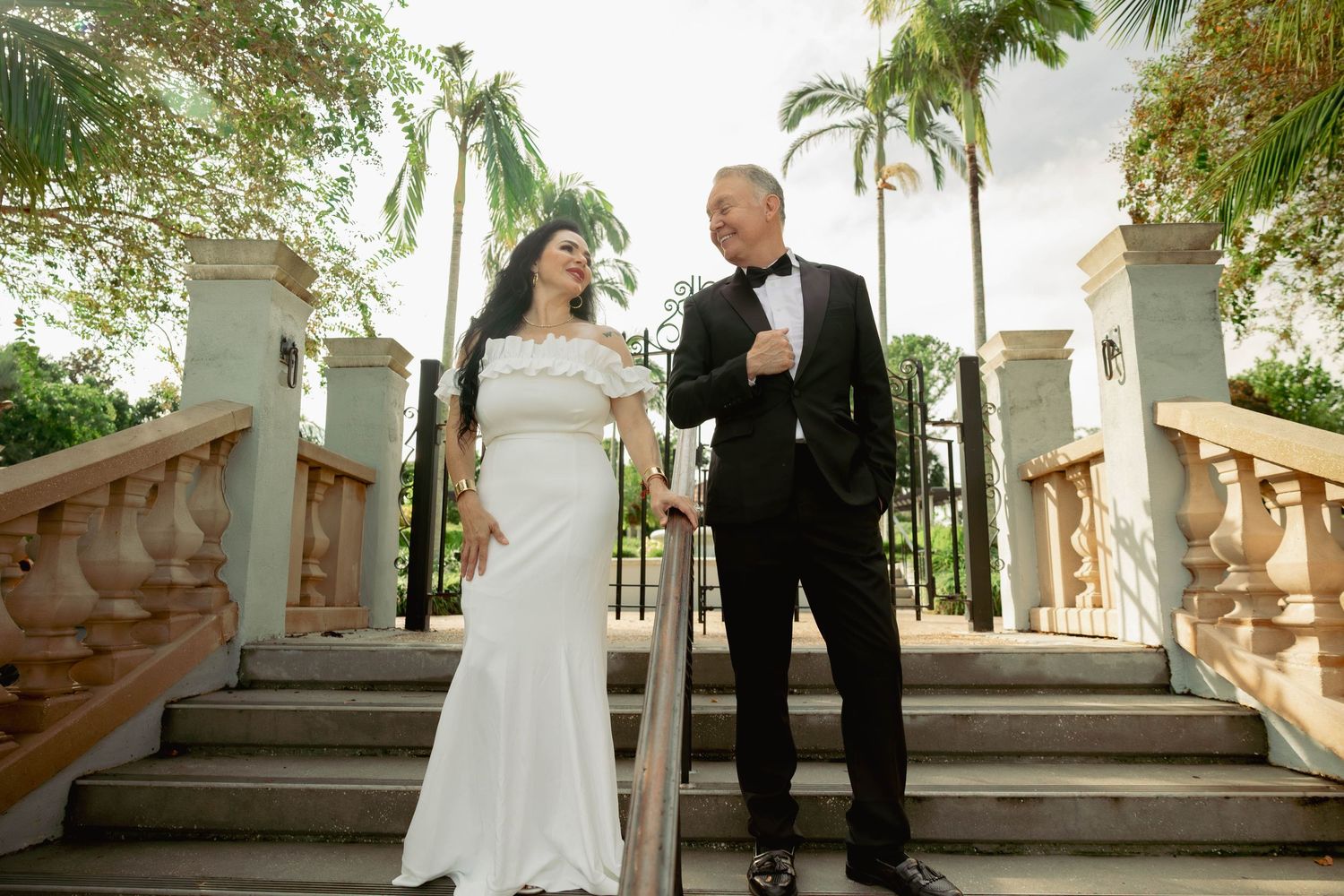 A bride and groom smile at each other on outdoor stairs surrounded by palm trees.