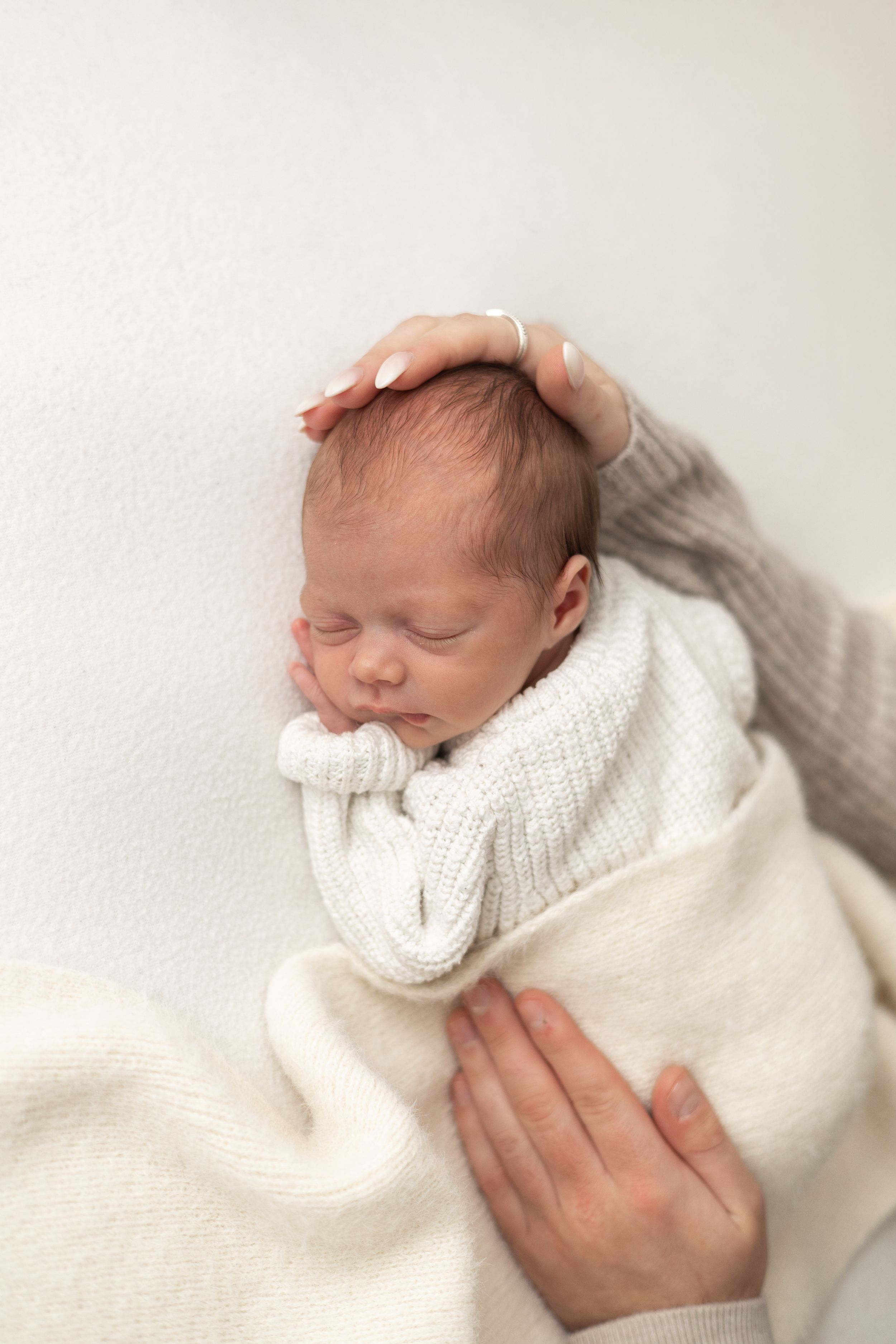 Newborn baby sleeping peacefully wrapped in white knit blanket while mother gently cradles with both hands Nottingham newborn photoshoot