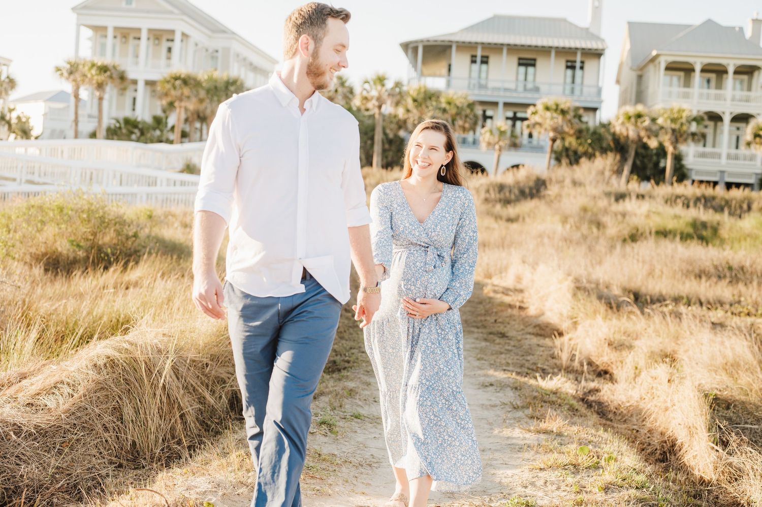 A couple walks hand in hand along a sandy beach path at sunset with coastal homes in the background.