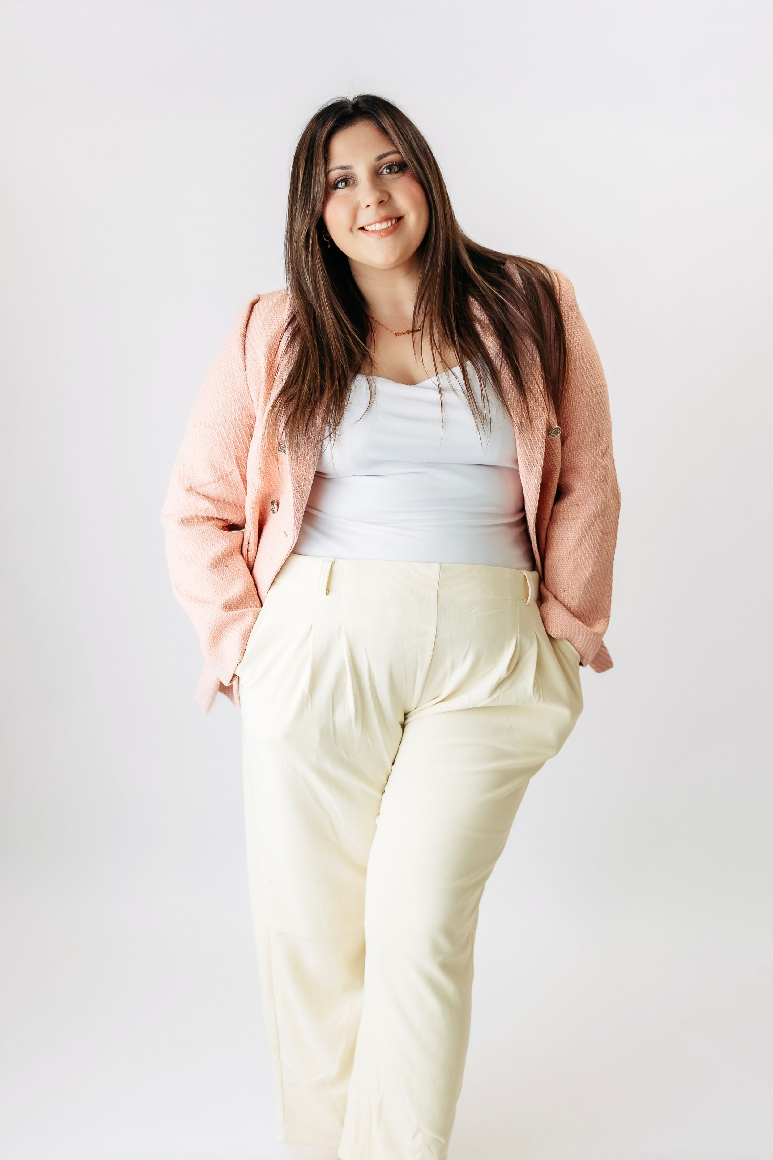Smiling brunette woman in pink blazer and cream pants posing on white background.