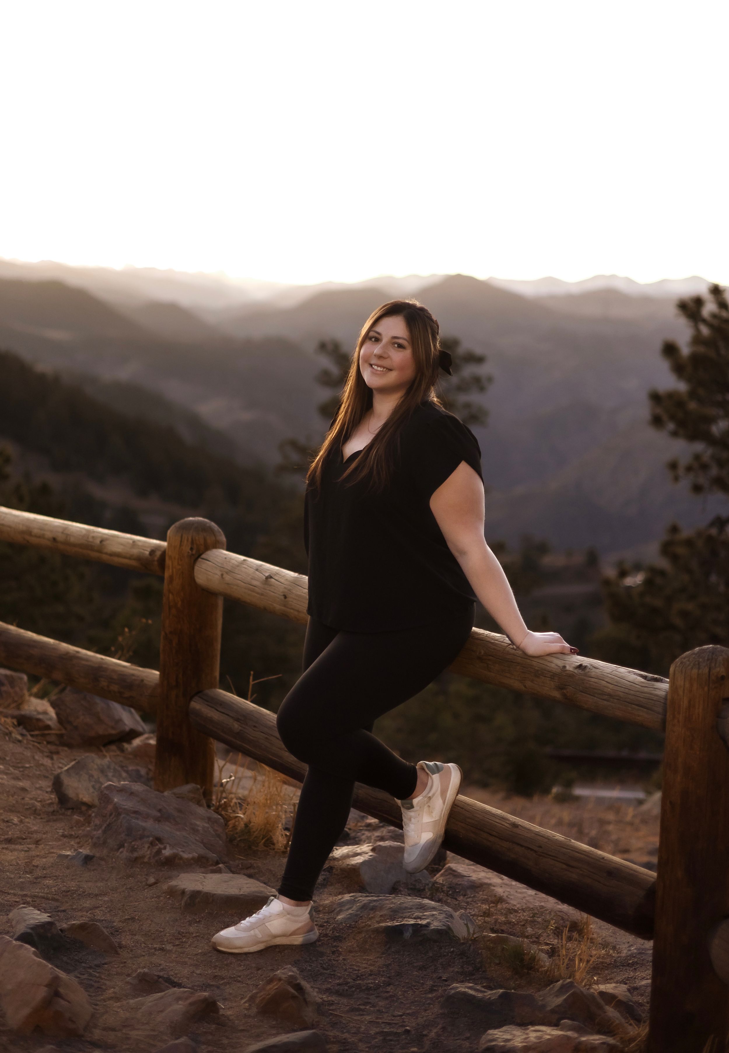 Smiling woman in black outfit leaning on wooden fence with mountain sunset backdrop.
