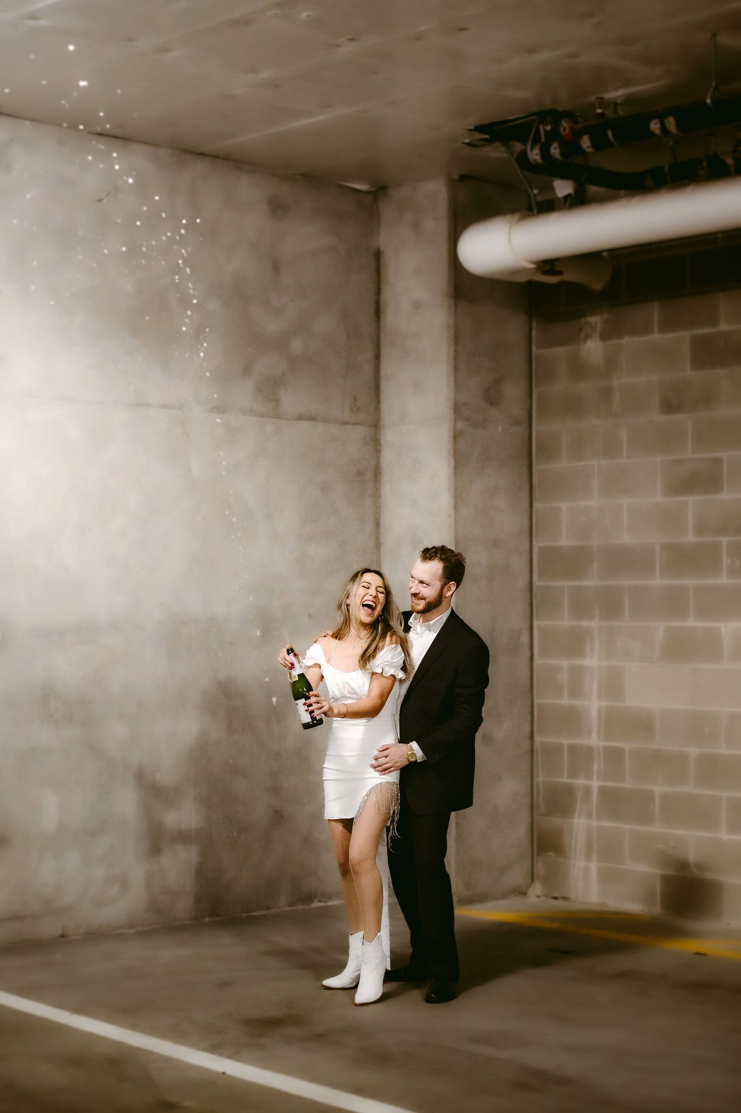 A couple in wedding attire poses playfully in a minimalist concrete parking garage with dramatic lighting.