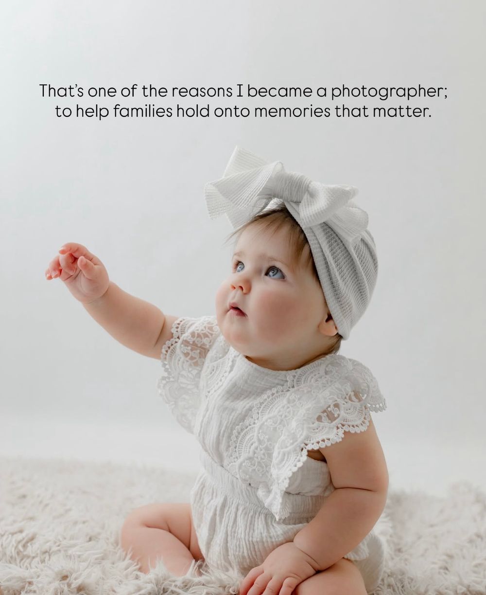 Baby wearing a delicate white lace dress and bonnet sits on a textured white blanket while reaching upward.