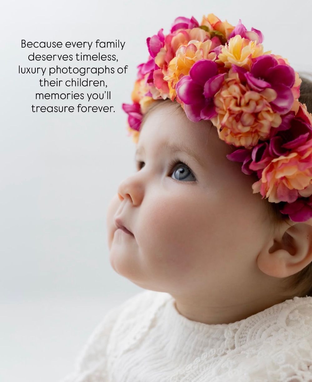 A baby wearing a white lace dress adorned with a vibrant floral crown of pink and peach blooms.