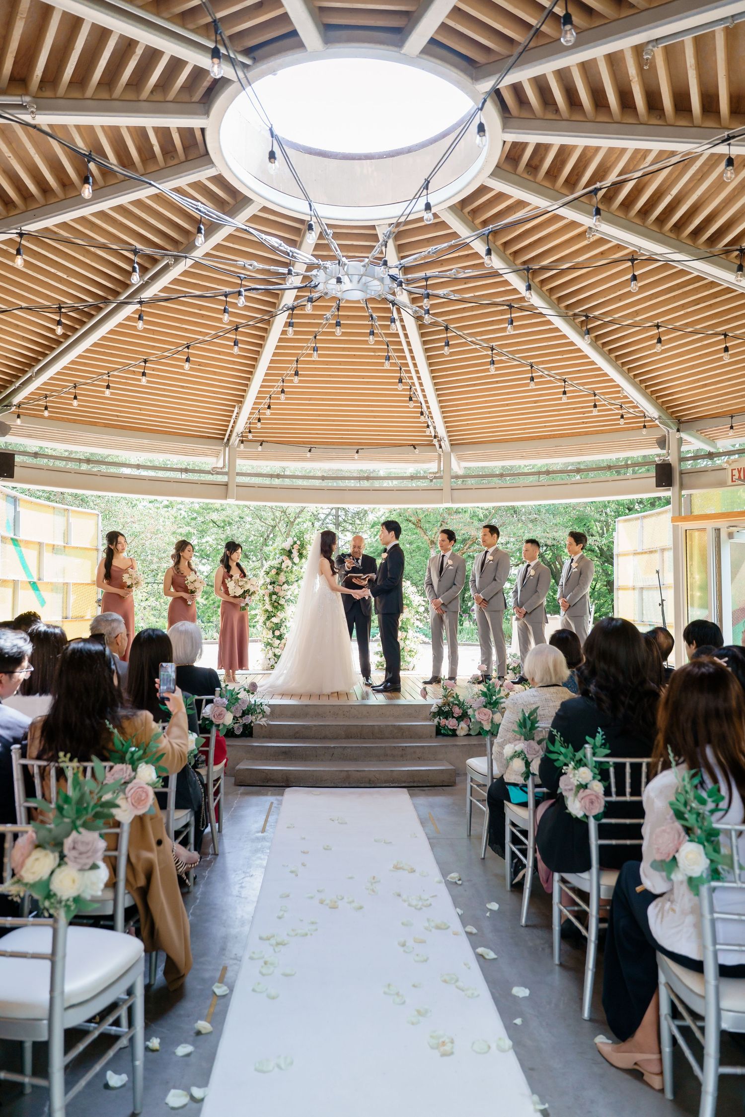 Panoramic view of an outdoor wedding ceremony venue with wooden gazebo and white chairs arranged in rows for guests.Queen Elizabeth Park Wed