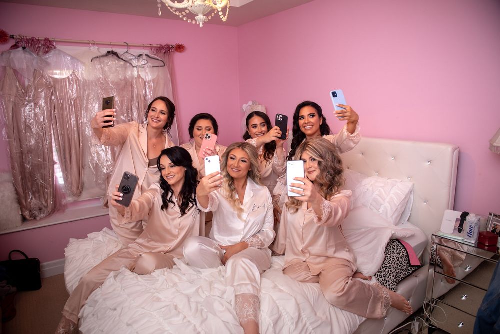 Bride and bridesmaids in pink satin robes taking selfies in a pink decorated bedroom.