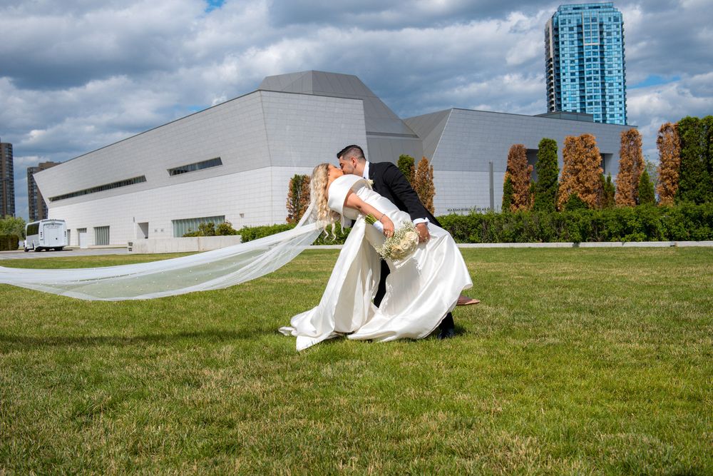 Groom dipping bride dramatically on green lawn with long flowing veil and modern building in background.