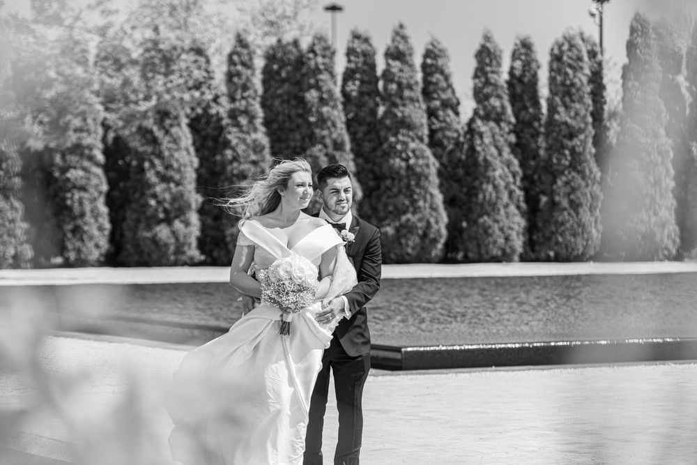 Black and white photo of bride and groom walking near a fountain surrounded by lush cypress trees.