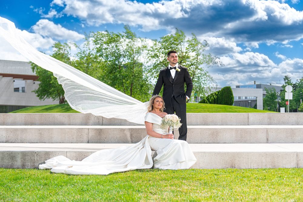 Bride and groom posing on green lawn with dramatic flowing veil under a bright blue cloudy sky.