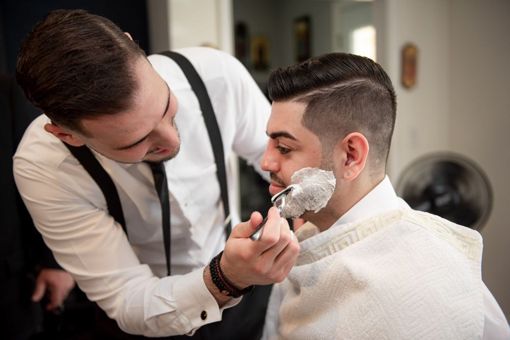 Barber applying shaving cream to groom's face during wedding day grooming preparation at barbershop.