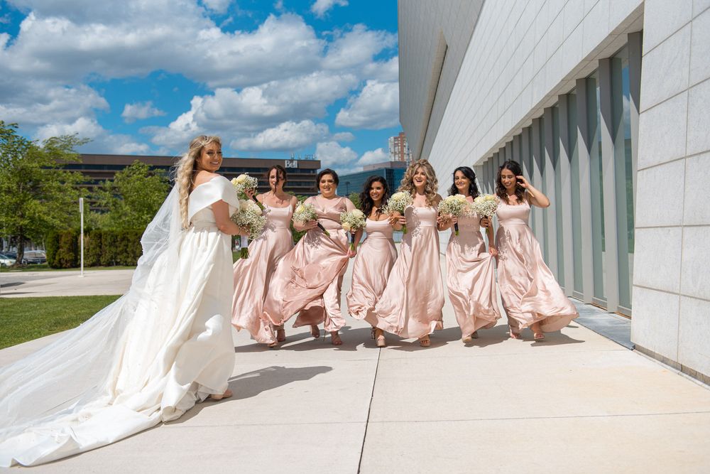 Bride in white gown leads bridesmaids in blush dresses walking joyfully outside modern building.