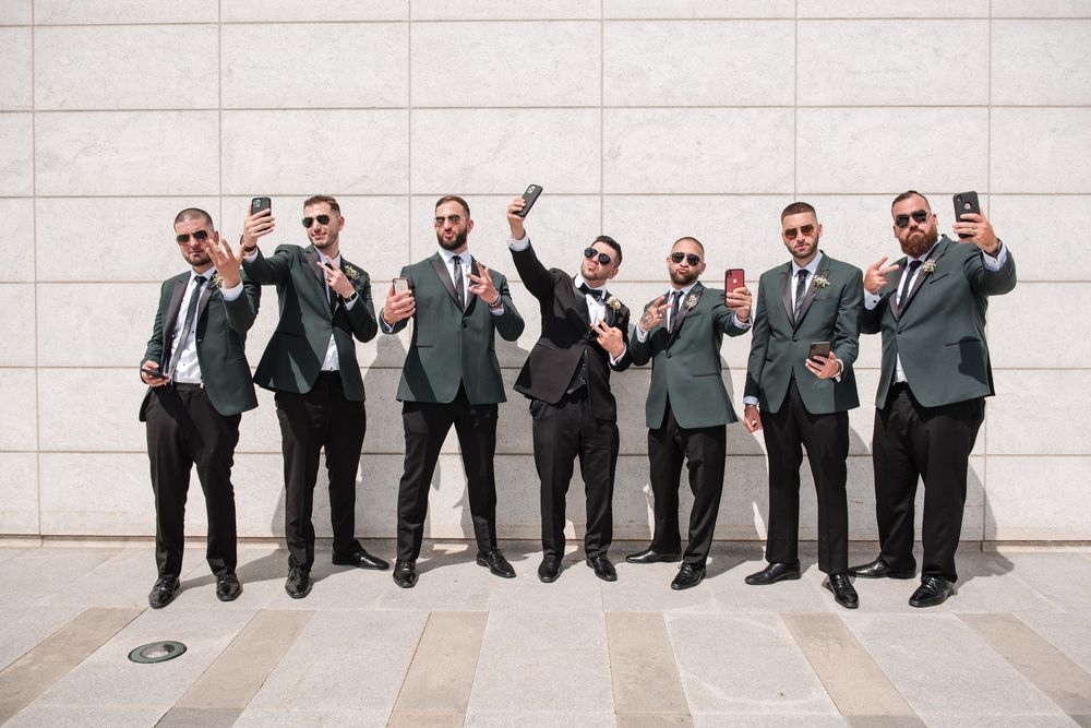 Groomsmen in green blazers take selfies while posing against a white marble wall outdoors.