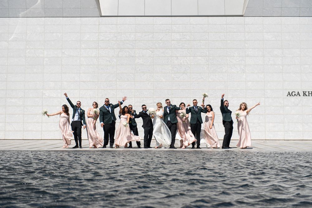 Wedding party in blush and black attire celebrate with raised arms near Aga Khan Museum waterfront.