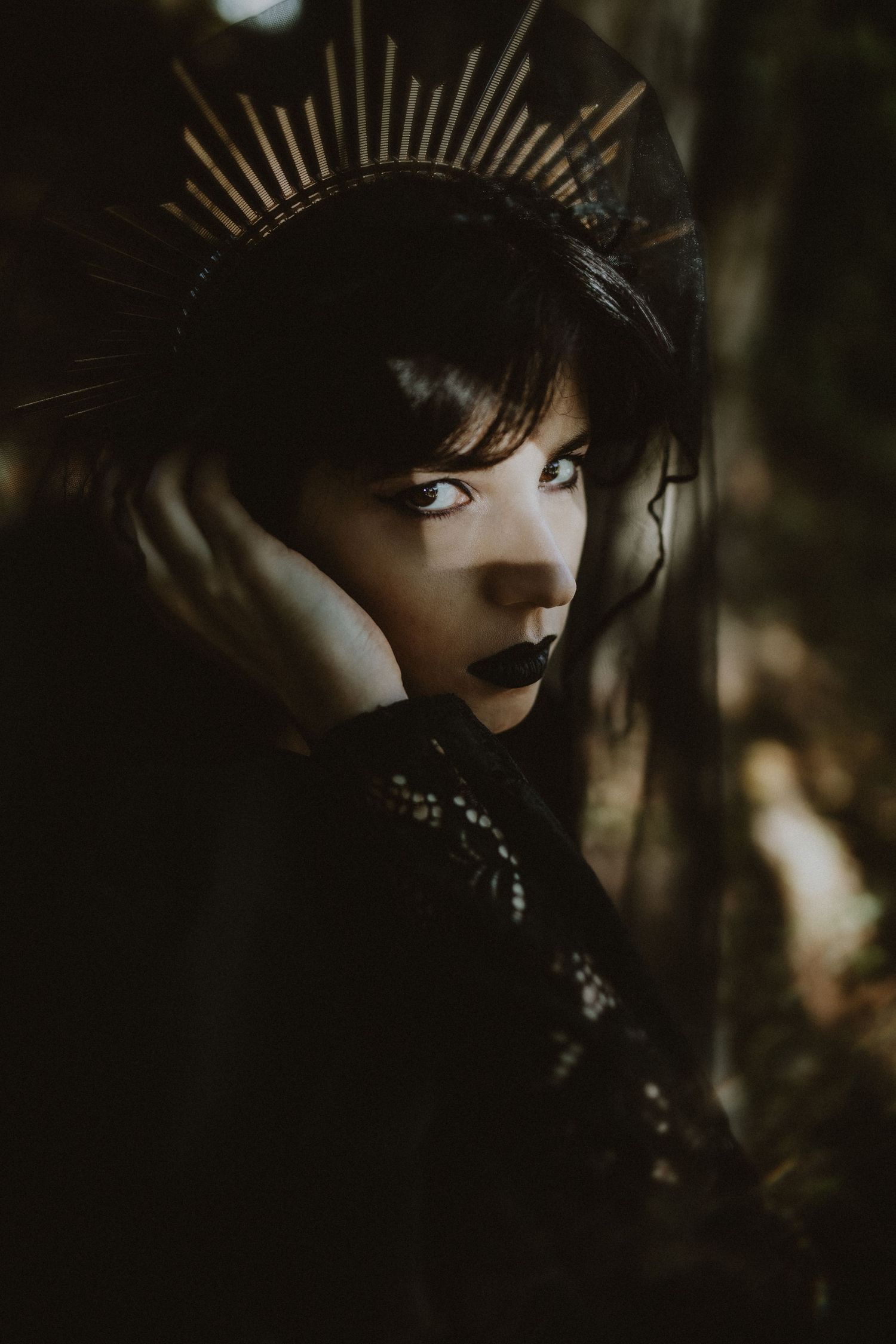 Woman in black gothic attire with gold crown headpiece and dark lipstick posing dramatically in a dark forest setting.