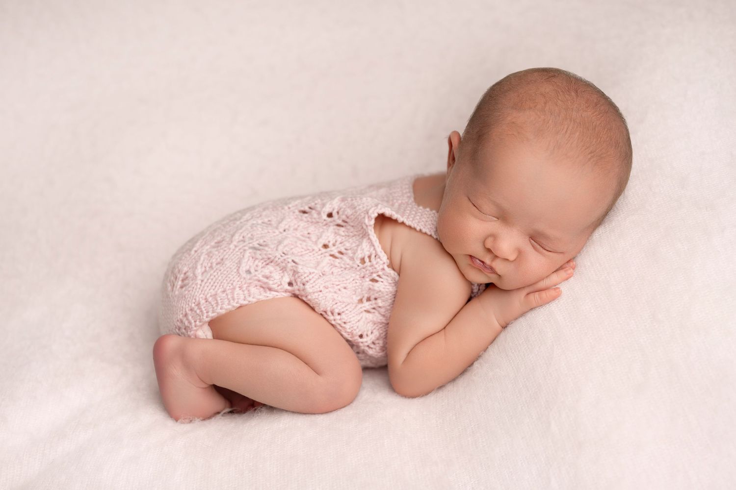 Sleeping newborn baby in pink knit romper on white fluffy blanket during her professional photography session.
