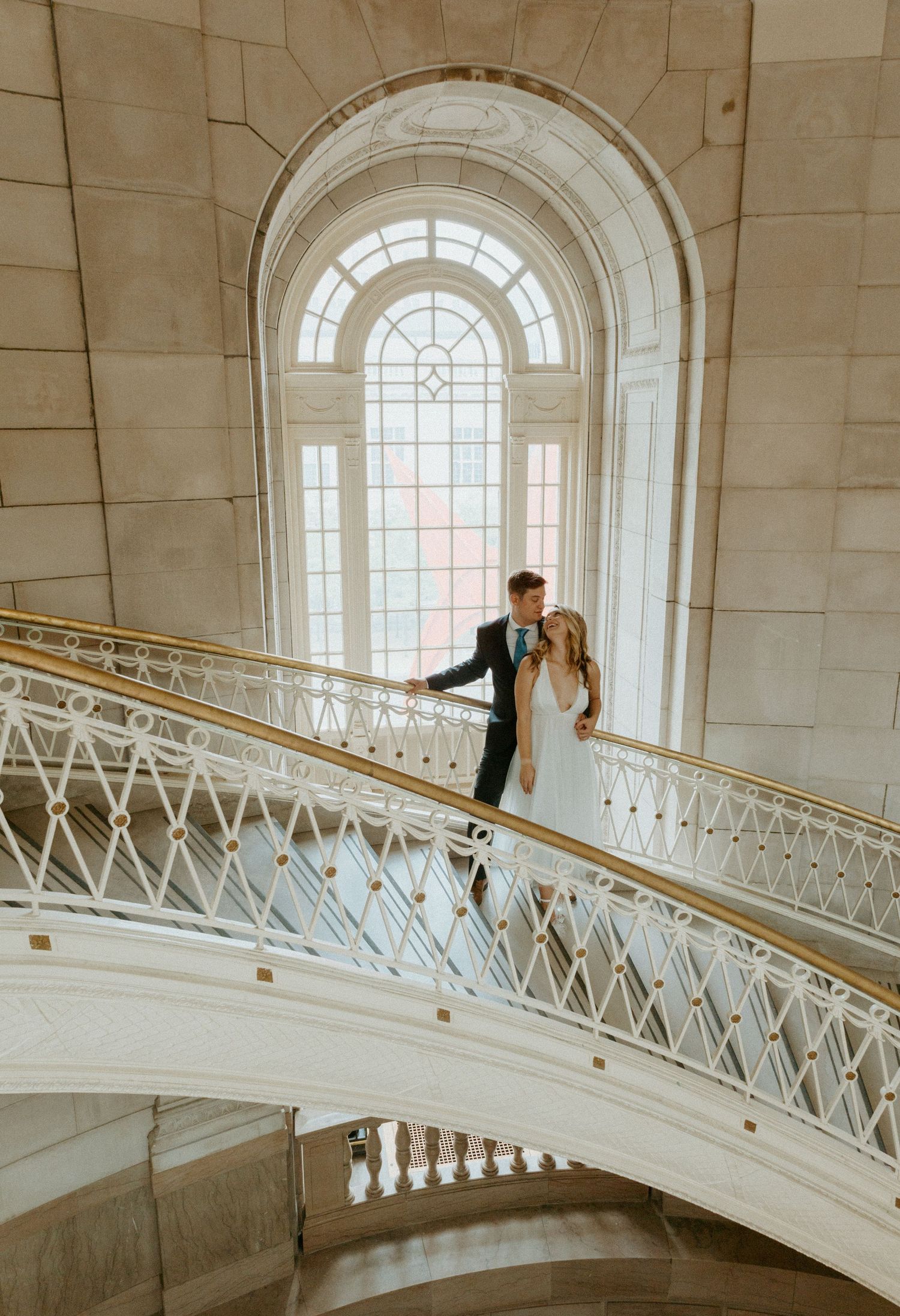 Bride and groom pose on elegant curved staircase with ornate white railing beneath arched window in grand venue.