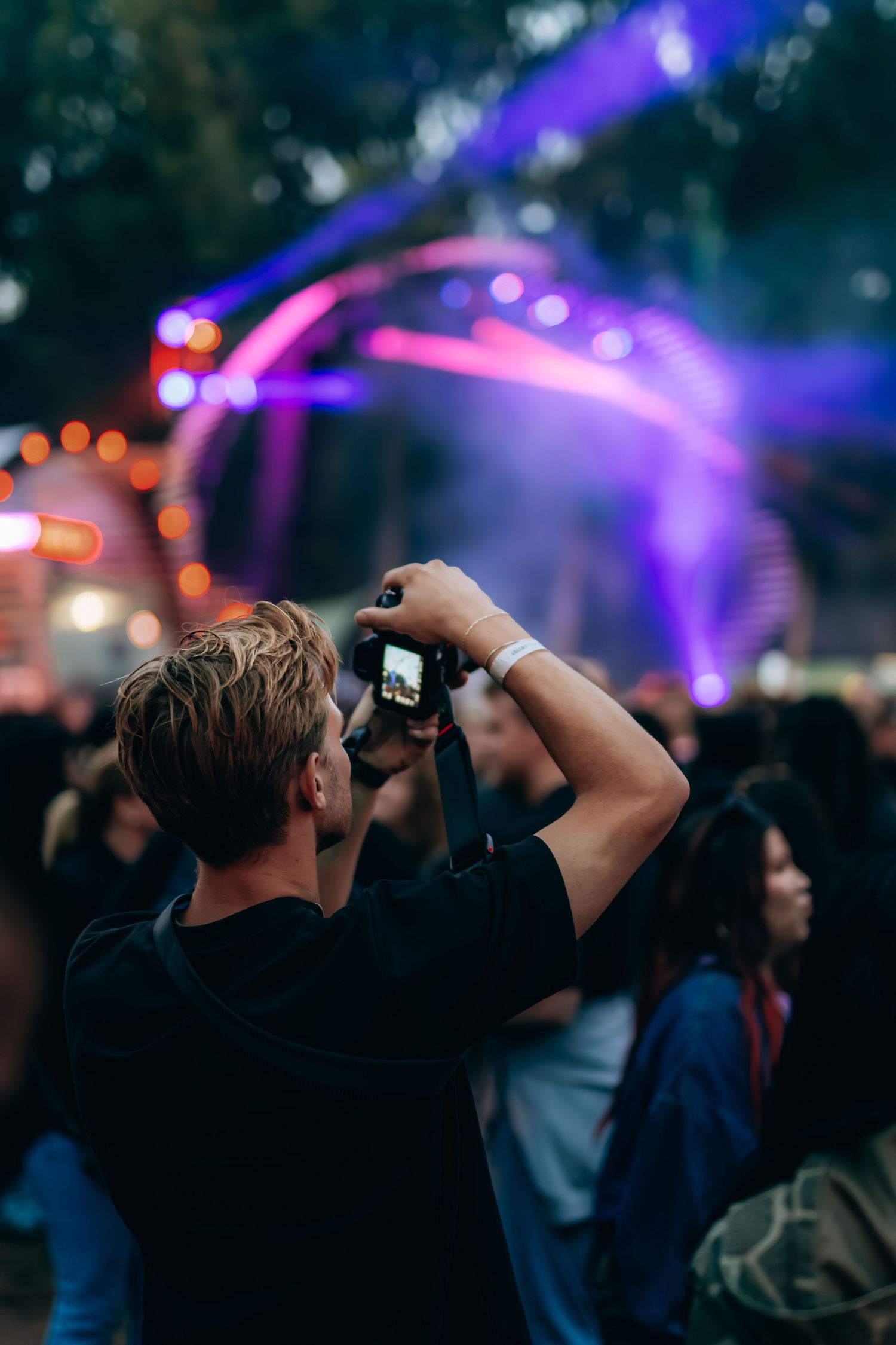 Man photographing colorful purple festival stage lights with a camera in a crowd.