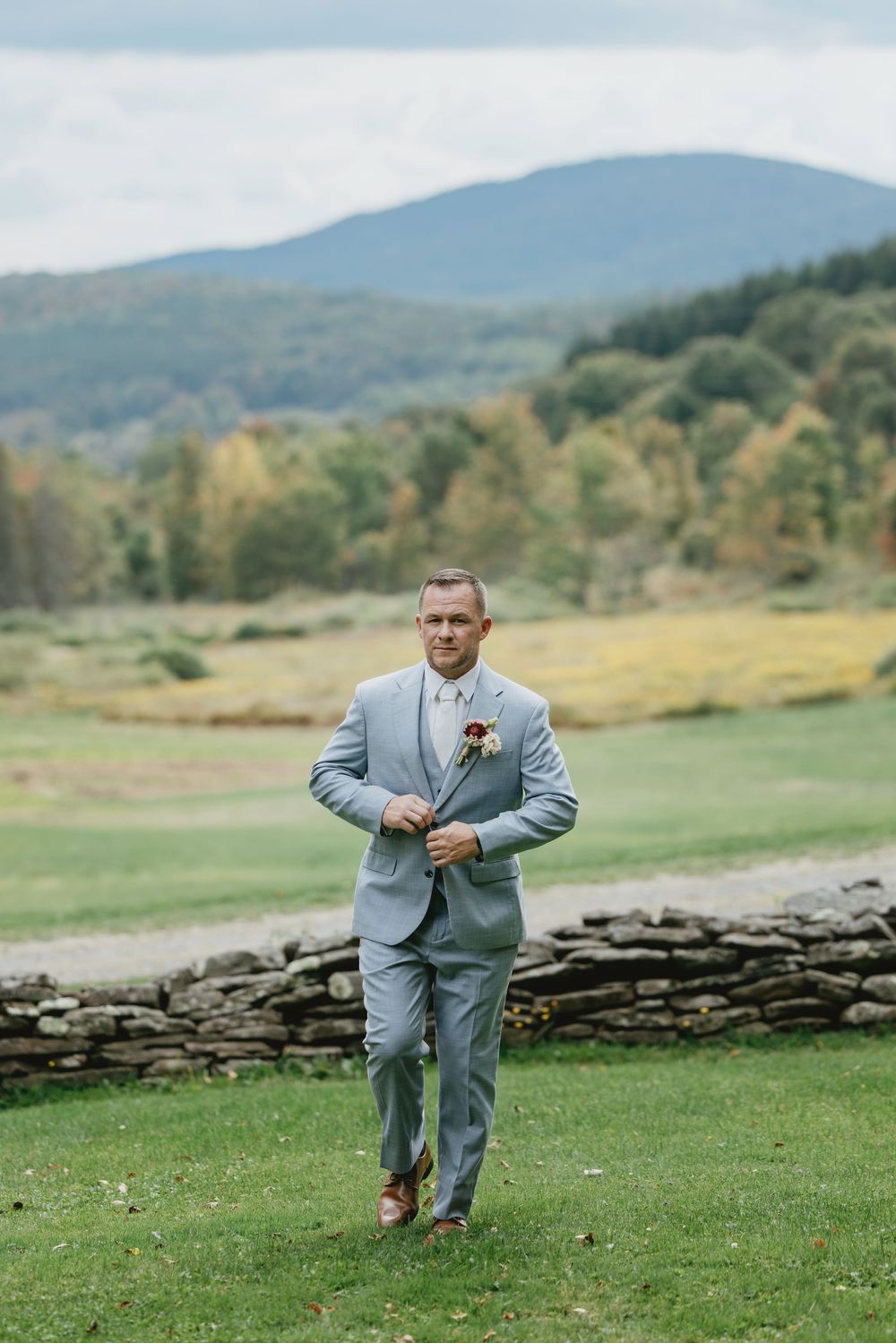 A groom in a light gray suit dashes across a scenic mountain meadow with stone walls.