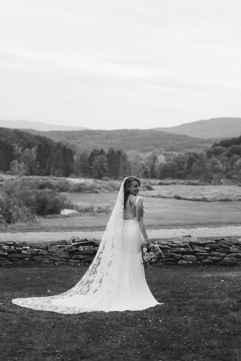 A bride in a flowing white wedding dress with long veil poses in a scenic mountain landscape setting.