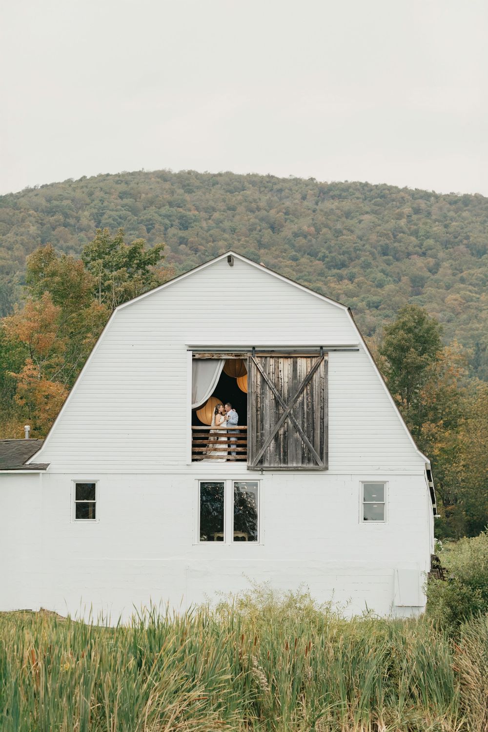 A white barn-style building with windows sits nestled against a forested hillside.